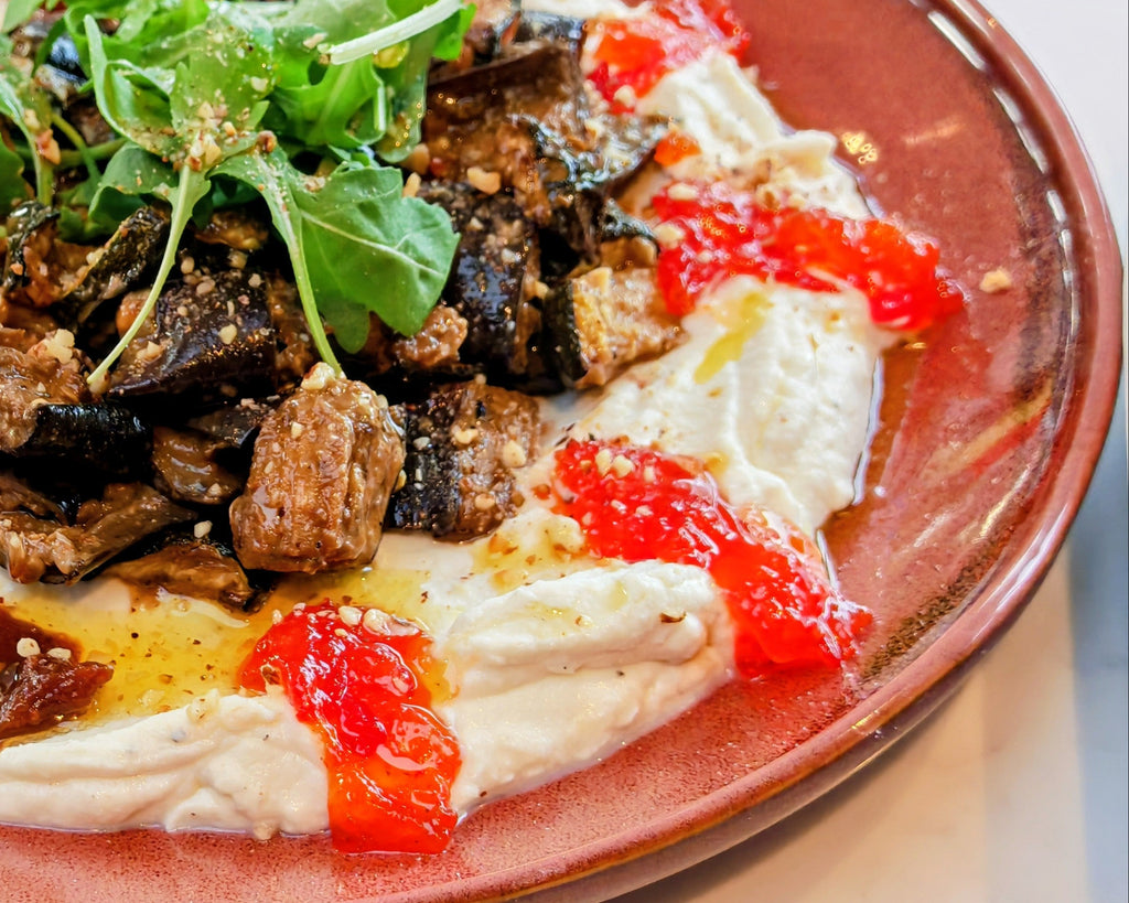 A close-up shot of a plated dish featuring creamy white hummus or baba ghanoush topped with sautéed eggplant, sun-dried tomatoes, and fresh green arugula, served on a terracotta-colored ceramic plate.