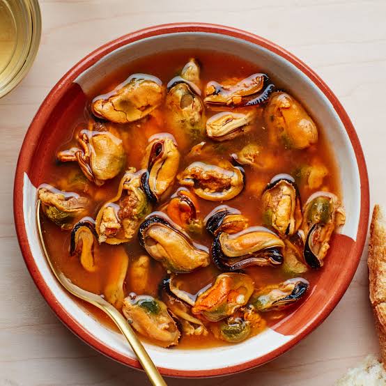 A top-down view of a bowl of mussels in a rich orange-brown sauce, served with a gold spoon on a light wood table.